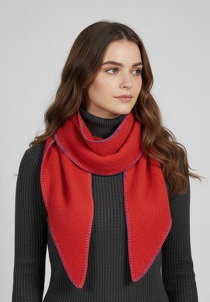 Junge Frau mit langen braunen Haaren trägt einen leuchtend roten Schal über einem dunkelgrauen, gerippten Rollkragenpullover und blickt leicht nach rechts.