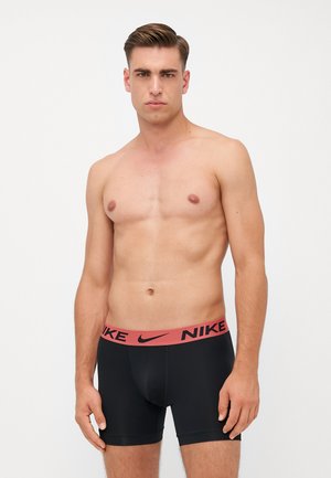 Schwarze Boxershorts mit einem breiten, rötlich-pinken Bund, der das Nike-Logo trägt. Der Stoff wirkt glatt und elastisch.