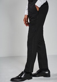 Pantalones negros ajustados con un corte recto, que cuentan con un bolsillo lateral y un sutil detalle de costura. Combinados con zapatos de vestir negros pulidos.