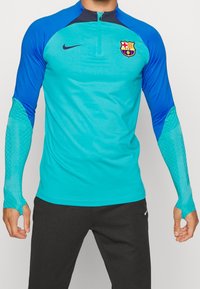 Nike träningsskjorta med en teal kropp, blå ärmar och dragkedja i nackpartiet. Inkluderar nätfack på ärmarna och FC Barcelona-logotypen på bröstet.