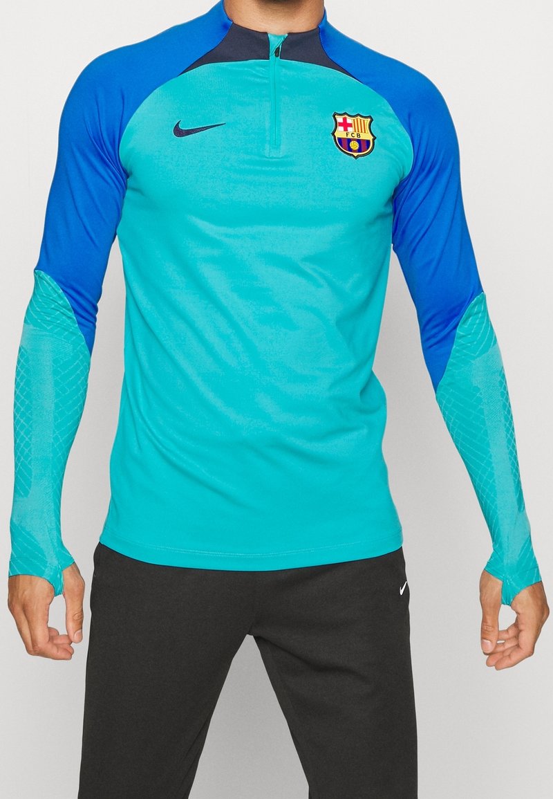 Nike träningsskjorta med en teal kropp, blå ärmar och dragkedja i nackpartiet. Inkluderar nätfack på ärmarna och FC Barcelona-logotypen på bröstet.