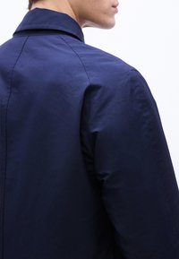 Άτομο που φοράει σκούρο μπλε σακάκι με γιακά, φαίνεται από  πίσω και ελαφρώς γυρισμένο, με φόντο ένα απλό ανοιχτό χρώμα.