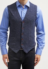 Marinblå texturerad väst med V-ringning, fem bruna knappar, två framfickor, bärs över en ljusblå skjorta, i kombination med mörka jeans.