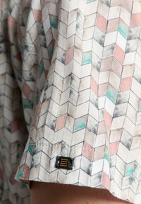 Kurzes Hemd mit geometrischem Muster in Pfirsich, Türkis und Grau, verziert mit einem schwarzen Etikett und gelben Akzenten am Saum.