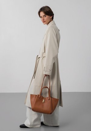 Vrouw draagt een lange beige trenchcoat en brede witte broek, met een grote bruine leren handtas tegen een eenvoudige grijze achtergrond.