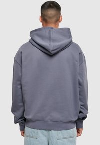 Mann med kort hår iført en løs grå hettegenser med hette ned og lyseblå jeans, sett bakfra mot en ensfarget bakgrunn.