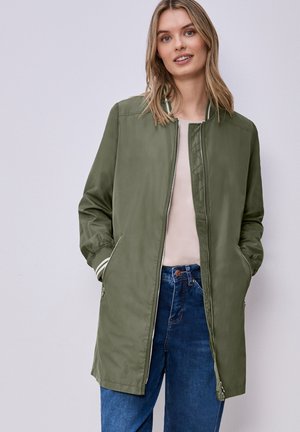 Donna bionda che indossa una lunga giacca verde oliva con zip, una maglia beige chiaro e jeans blu, in piedi con le mani nelle tasche davanti a uno sfondo semplice.