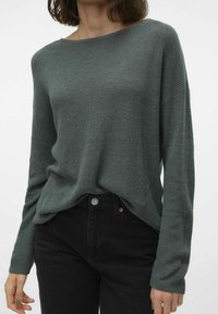 Grüner, gestrickter Pullover mit langen Ärmeln, weitem Ausschnitt und lockerer Passform, kombiniert mit schwarzen Jeans. Weiche Textur und geripptes Muster.