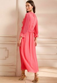 Abito lungo in corallo con maniche lunghe plissettate, vita cintrata e dettagli a volants; abbinato a scarpe con tacco gold su un pavimento in legno.