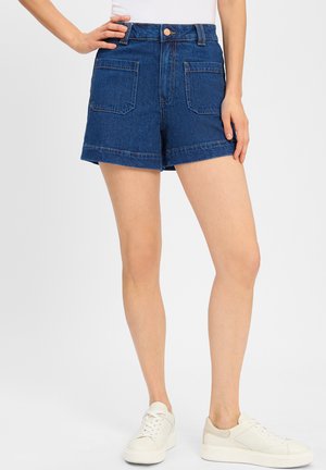 Denim shorts med høj talje, blå farve, foldet kant og to baglommer, parret med hvide sneakers med en lys tan-accent.