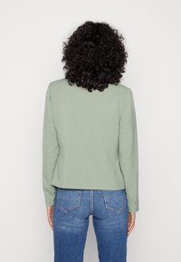 Blazer verde, ajustado, con mangas largas, corte recto y textura suave. Combinado con jeans de mezclilla azules. Vista trasera mostrada.