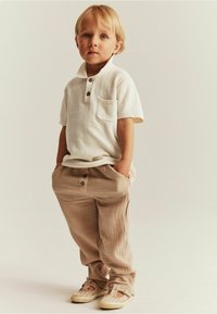 Kurzärmliges, cremefarbenes Poloshirt mit einer Tasche und zwei Knöpfen; hellbeige, strukturierte Hose mit Knöpfen; weiche Schuhe mit offenes Design.