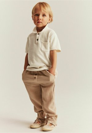 Kortärmad, krämfärgad polo med ficka och två knappar; ljusbeige, texturerade byxor med knappar; mjuka skor med öppen design.