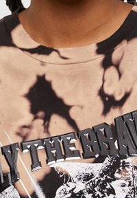 Grafický tričko s tie-dye vzorem v tan a hnědé barvě, opatřené velkým lesklým černým textem "JEN TI NEJODVAŽNĚJŠÍ" na hrudi.