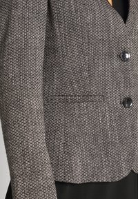 Rakennepintainen mustavalkoinen bleiseri, jossa on lyhyt malli, yksi tasku, kaksi pyöreää mustaa nappia ja hienovaraisesti kudottu kuvio.