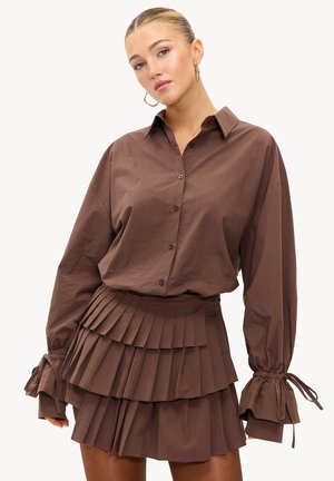 Chemise marron à manches longues avec boutons, associée à une mini-jupe plissée assortie. La chemise comporte des liens aux poignets et une coupe décontractée ; la jupe présente des plis superposés.