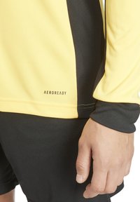 Gelbes Sportshirt mit schwarzen Akzenten, gerippten Bündchen und dem "AEROREADY"-Logo nahe dem Saum. Glattes, feuchtigkeitsableitendes Material.