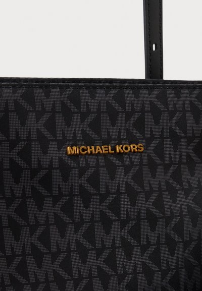 Близък план на черна чанта тоут Michael Kors с повтарящ се мотив на логото, златист надпис на марката и черна презрамка.
