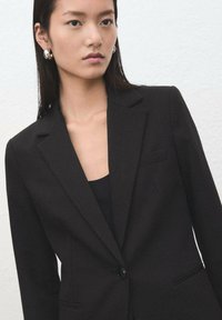 Czarna marynarka wykonana z dopasowanej tkaniny, z zapięciem na jeden guzik, klapami z wycięciem typu notch oraz dwoma bocznymi kieszeniami. Elegancki, dopasowany krój.