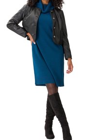 Giacca cropped in pelle nera, vestito in maglia teal con scollo a cowl, stivali neri alti fino al ginocchio in camoscio. Focus su tessuti testurizzati e contrasto di colori.