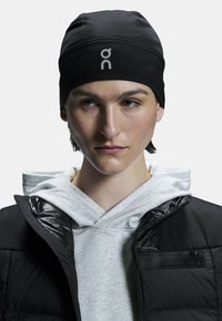 Bonnet noir avec logo, porté sur un hoodie gris et une doudoune noire. Tissu lisse avec un design ajusté, détails de couture sans couture.