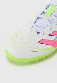 Chaussure de soccer blanche avec une semelle extérieure vert fluo, des accents roses et un dessus texturé présentant un motif. Les lacets sont blancs avec un détail en maille verte.