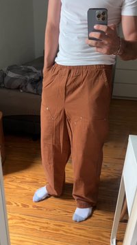 Uomo che indossa pantaloni cargo marroni, calzini bianchi e una maglietta bianca mentre si fa un selfie allo specchio in una camera da letto con pavimento in legno e un letto.
