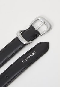 Svart läderbälte med silverfärgad metallspänne, med en slät yta och sömdetaljer. "Calvin Klein" tryckt i vitt på remmen.