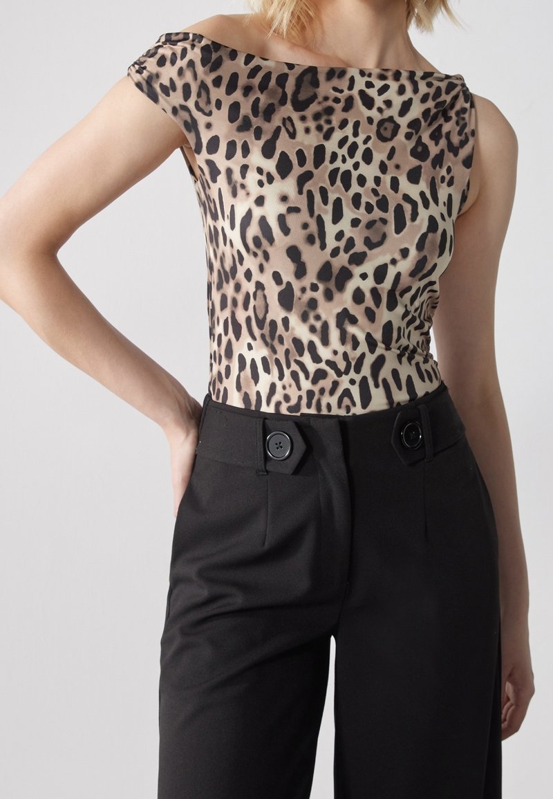 Femme portant un haut sans manches à imprimé léopard, rentré dans un pantalon noir taille haute avec de gros boutons à la taille.