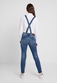 Denimoverall med justerbara blå remmar, figursydd design, ljus blekning på benen, och en brun läderpatch i midjan. Vit skjorta under.