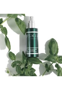 Grøn metallisk sprayflaske med sølvdetaljer, mærket "OSKIA LONDON CITYLIFE FACIAL MIST", omkranset af friske grønne basilikumblade.