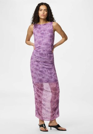 Kvinde med krøllet mørkt hår iført en ærmeløs lilla blomster maxi-kjole og sorte åbne hæle, stående med hænderne på hoften.