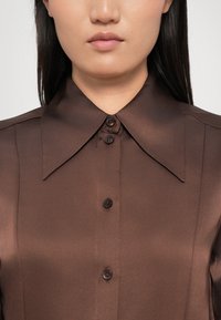 Cămașă din satin maro cu un guler clasic lat, având un sistem de închidere cu nasturi pe față și o textură netedă. Cinci nasturi negri sunt vizibili pe placket.