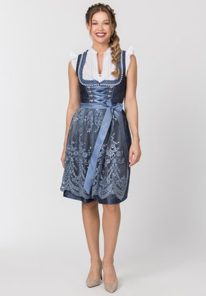 Frau mit geflochtenem Haar, die ein dunkelblaues Dirndlkleid mit Spitzen-Schürze und hellblauem Taillenband trägt, steht und lächelt.