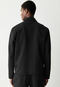 Chaqueta negra con una textura de tela suave, que presenta un cuello clásico, costuras invisibles y un diseño minimalista. Vista posterior mostrada.