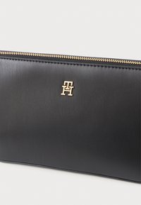 Svart lærveske med gullglidelås og liten gull "TH" logo sentrert under glidelåsen på ensfarget bakgrunn.