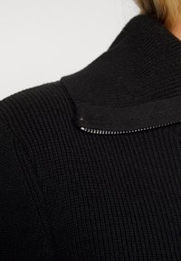 Czarna prążkowana tkanina o teksturowanej powierzchni z detalem kołnierza na suwak. Materiał wydaje się miękki i nieco rozciągliwy.