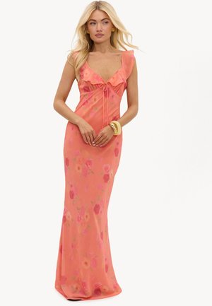Femme aux longs cheveux blonds portant une robe longue corail rose à motif floral avec un col à volants, debout, les mains jointes.