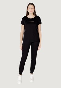 T-shirt in cotone nero con colletto rotondo e maniche corte, abbinata a pantaloni sportivi neri. Le sneakers bianche creano un contrasto. Design semplice e raffinato.