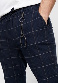 Námořnické kostkované kalhoty s pasem na stažení, zdobené stříbrným řetízkem a kovovým kruhem. Tkanina má texturu s mřížkovým vzorem a bílým prošitím.