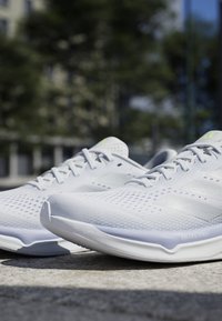 Een paar witte atletische hardloopschoenen met een mesh-ontwerp, buiten geplaatst op beton met vervaagde bomen en gebouwen op de achtergrond.