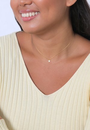 Collier doré avec un petit pendentif rond, porté avec un top à col en v côtelé de couleur jaune clair, mettant en valeur le cou et la zone de la clavicule.