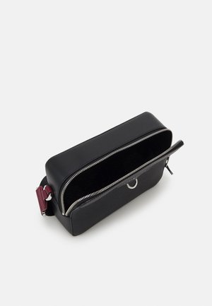 Zwarte rechthoekige crossbodytas met gedeeltelijk open zilveren rits, met een bordeauxrode riem bevestiging op een witte achtergrond.