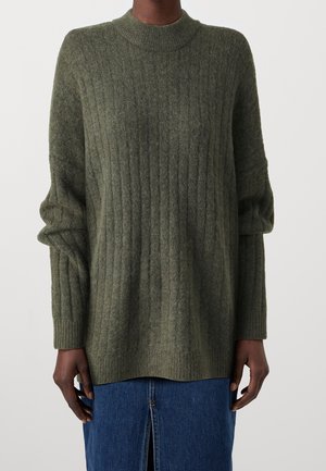 Personne portant un pull ample côtelé vert olive à manches longues et une jupe en jean bleue avec une fente à l'avant.