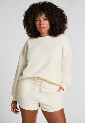 Cremfarbener, weicher Baumwollpullover mit überschnittenen Schultern und Rundhalsausschnitt, kombiniert mit passendem Shorts mit Kordelzug. Glatte Textur.