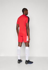 Rød sportsdrakt med svarte detaljer, kombinert med matchende røde shorts. Hvite knelange sokker og svarte fotballsko fullfører antrekket.