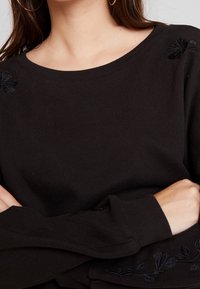 Jersey negro con un escote amplio y acentos florales bordados en las mangas. Textura de punto suave con mangas largas y de corte holgado.