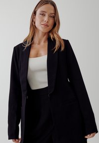 Černá dressovaná sako s klopami, jedním předním knoflíkem a kapsami. Nosené přes krémový vroubkovaný top s dlouhými rukávy.
