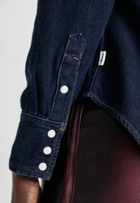 Jeansowa koszula z ciemnoniebieskimi rękawami i białymi guzikami. Posiada pomarańczowe przeszycia oraz mankiety zapinane na guzik, co pozwala na regulację dopasowania.