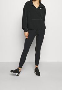 Sort Nike zip-up jakke med snoretræk i kanten, parret med sorte leggings og sorte sportssko. Glat stof med minimale designdetaljer.